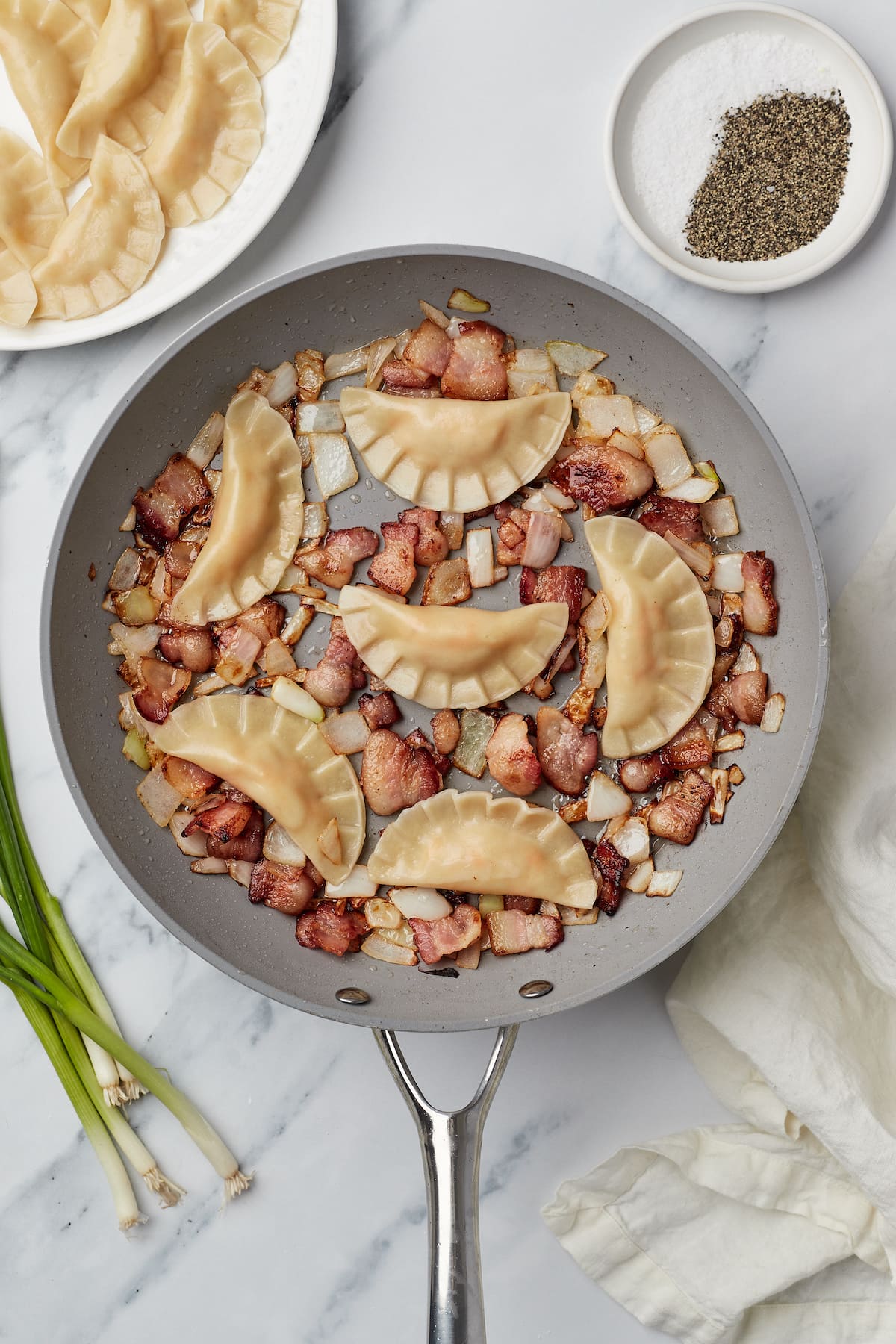 A skillet with bacon, onions, and semi-circular dumplings made with wonton wrappers.