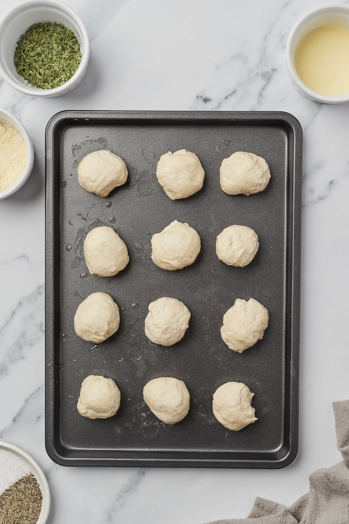 Small pieces of unbaked yeast dough on a baking sheet. Ramekins of parsley, melted butter, salt and pepper, and Parmesan are arranged around the baking sheet.