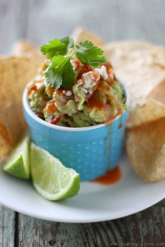 Buffalo Guacamole in a blue ceramic container topped with cilantro all on a larger white plate with chips and lime wedges
