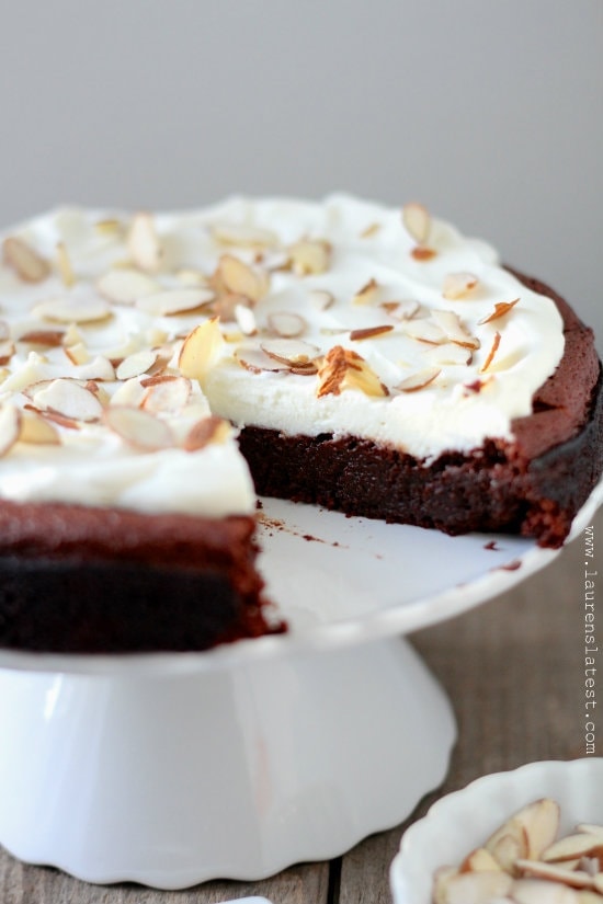 Flourless Chocolate Cake with a missing slice, topped with with Almond Whipped Cream and almonds all on a white cake platter