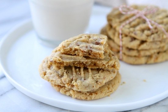 a stack of peanut butter toffee cookies on a white plate. the top cookie is broken and there is another stack in the background with a glass of milk