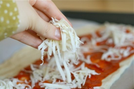 sprinkling cheese on top of pizza sauce on rolled out pizza dough
