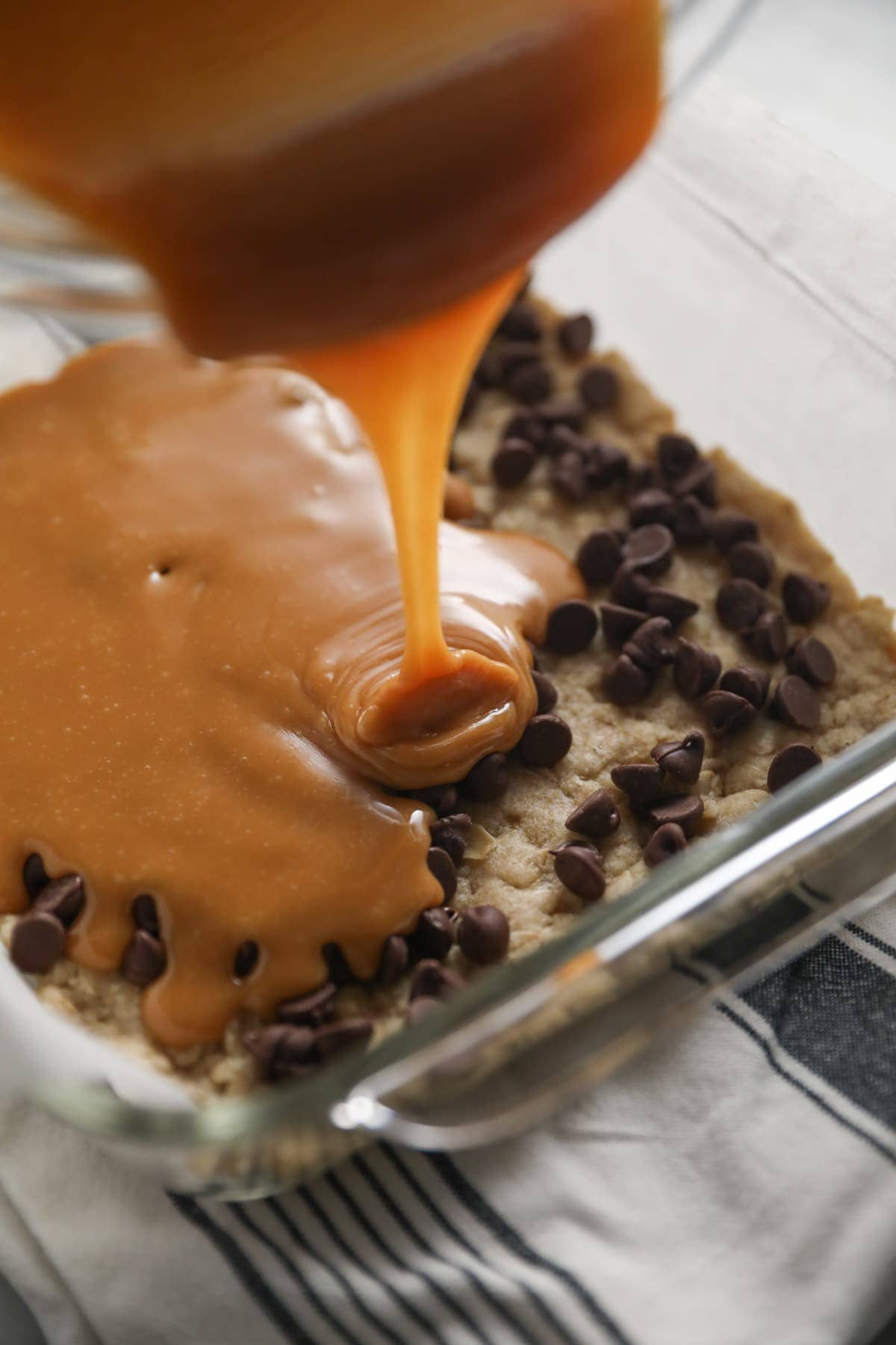 pouring melted caramel over chocolate chips and oatmeal cookie dough base