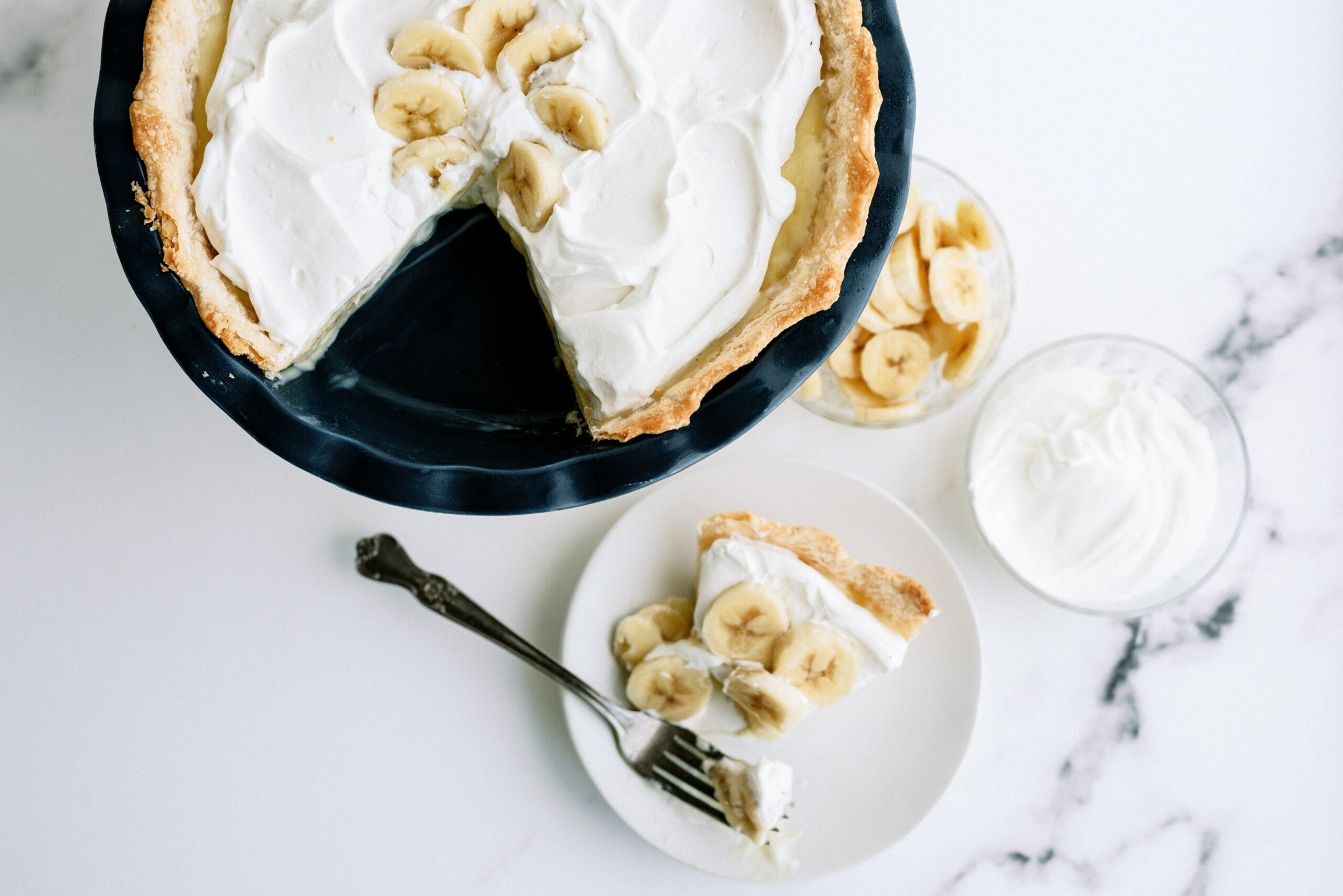 banana cream pie with a slice taken out of it. the slice is on a white plate next to the whole pie