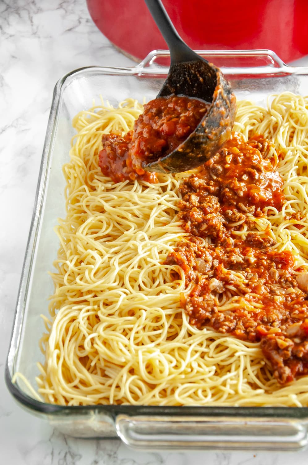 spreading red sauce over cooked spaghetti in a glass baking dish