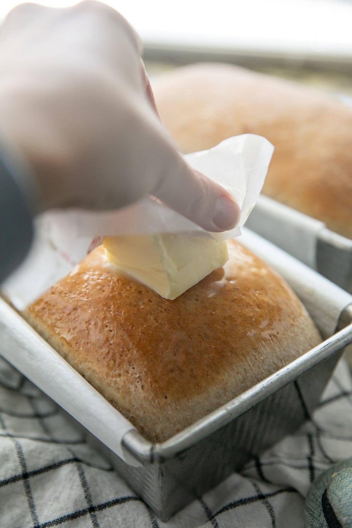 a hand rubbing a stick of butter on a freshly baked loaf of bread still in the pan