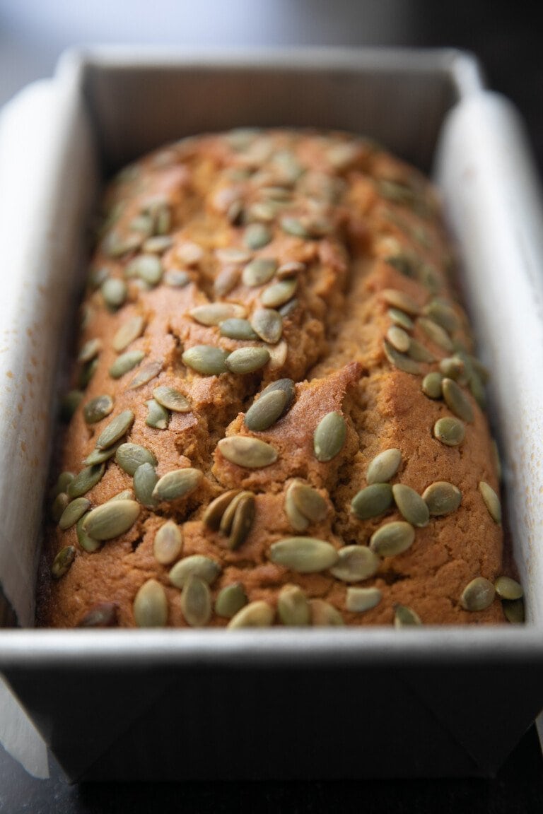 baked pumpkin bread in a parchment paper lined loaf pan topped with pepitas