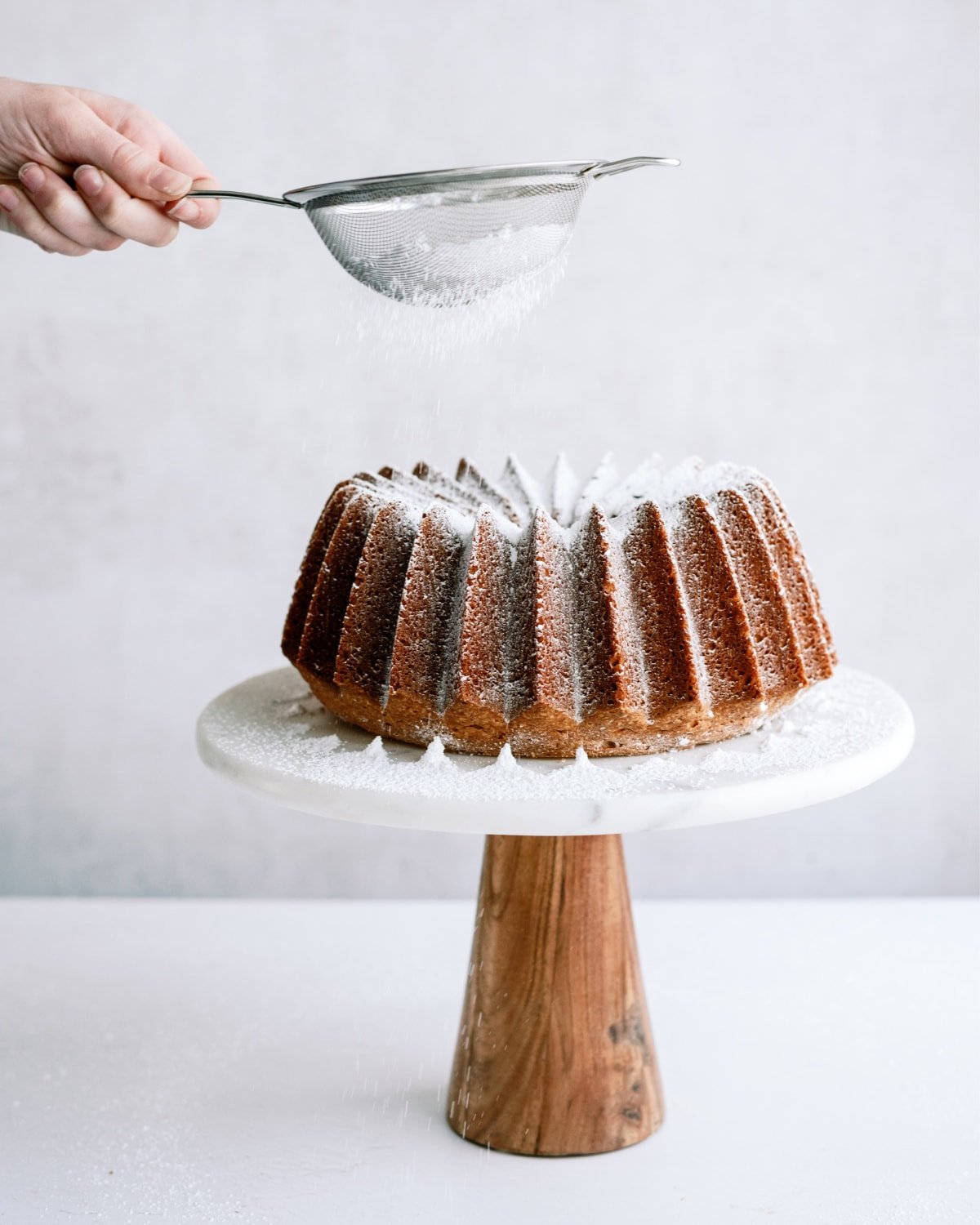 vanilla bundt cake on platter with woman dusting sugar