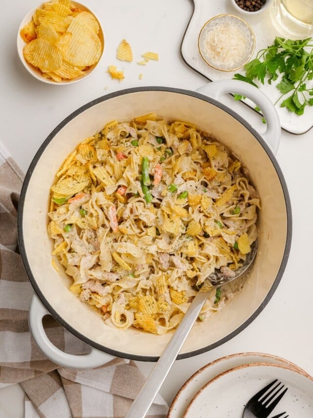 A pot of creamy pasta with vegetables and crumbled potato chips on top. A serving spoon rests inside. A small bowl of potato chips, a fork, a plate, and garnishes are nearby.