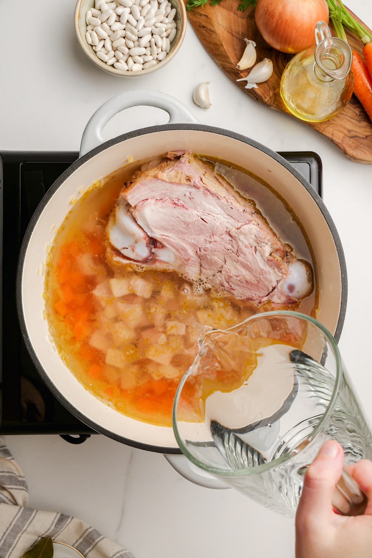 woman's hand pouring water into a pot with ham and bean soup ingredients