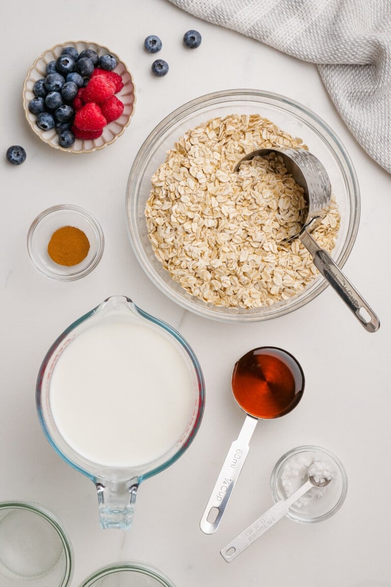 For overnight oats, gather a bowl of oats with a scoop, milk in a measuring cup, a small dish of cinnamon, honey in a measuring cup, salt, and a bowl of raspberries and blueberries. Simply mix and refrigerate for a nourishing breakfast delight.