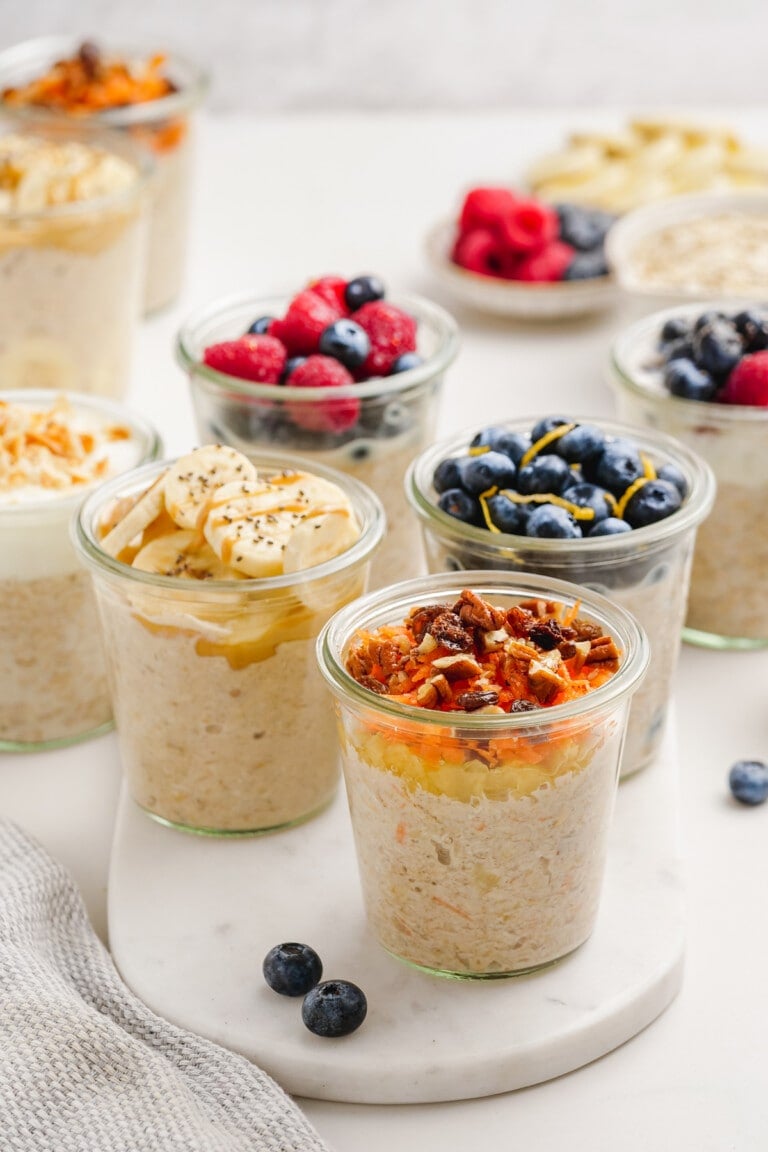 Various jars of overnight oats topped with fruits, nuts, and seeds are arranged on a white surface. Blueberries and raspberries are scattered around, enhancing the vibrant display of these nutritious morning delights.