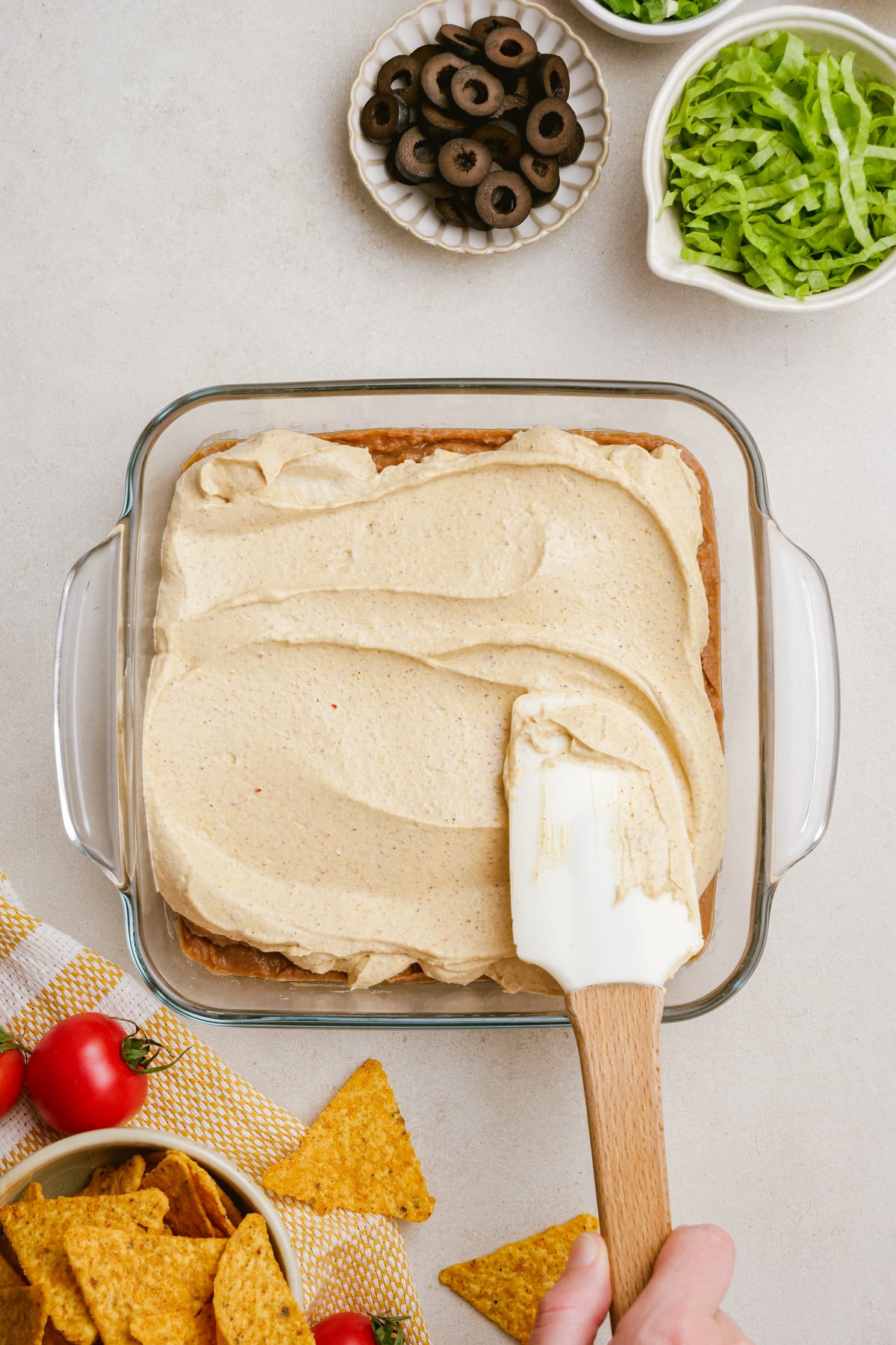 woman's hand spreading cream cheese mixture on top of refried beans in casserole dish with a rubber spatula