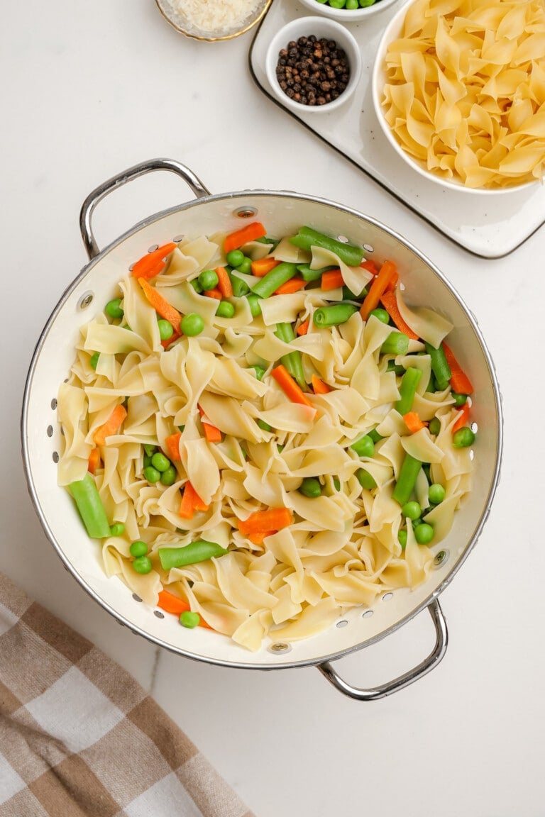 A colander filled with cooked pasta, green beans, peas, and sliced carrots evokes the essence of a vibrant Tuna Noodle Casserole. In the background, dishes brim with more pasta alongside small bowls of black pepper and grated cheese.