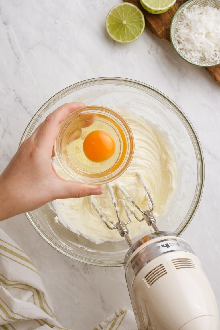 woman's hand adding egg to cream cheese and sugar in a bowl