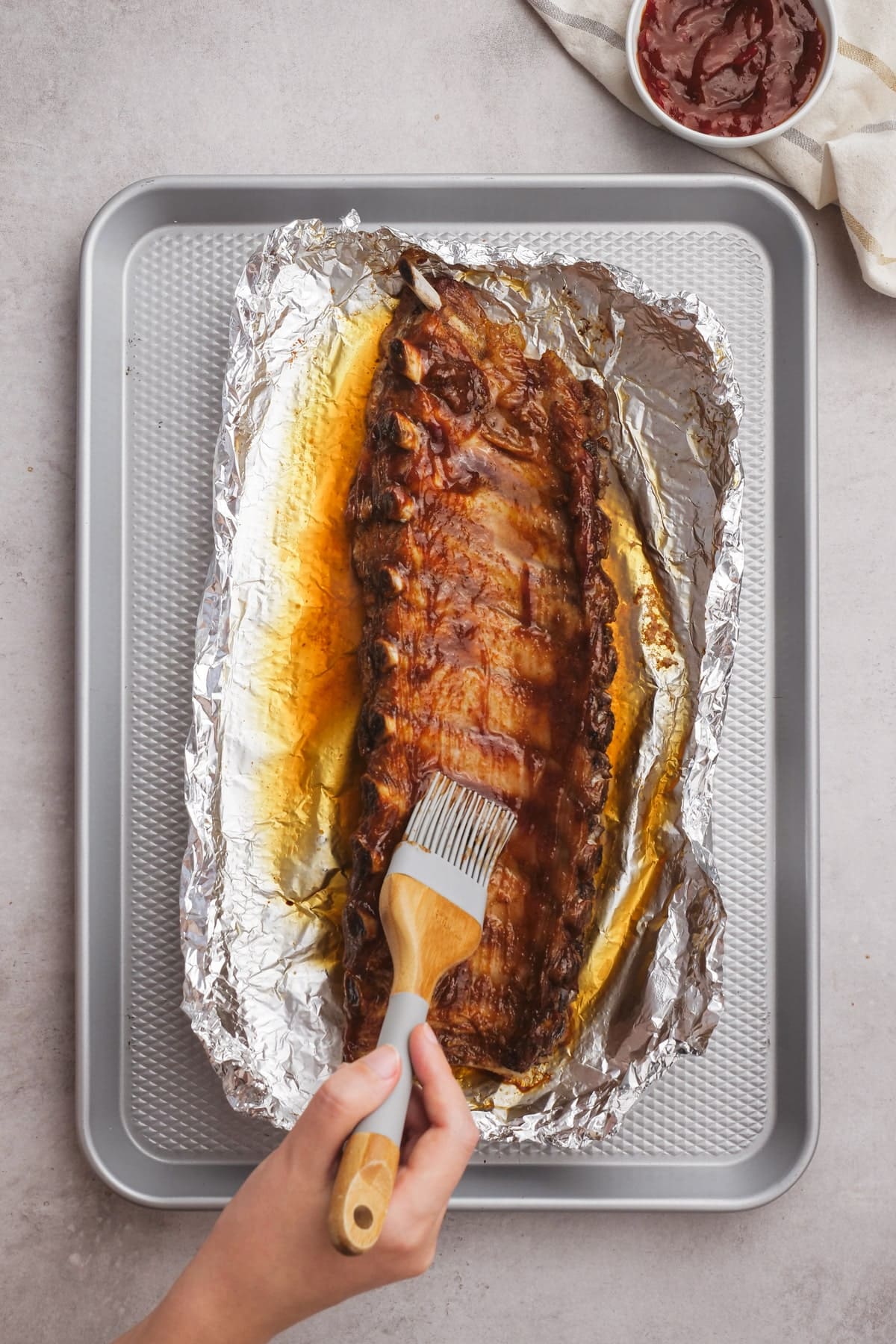 woman's hand brushing bbq sauce on ribs in foil on baking sheet