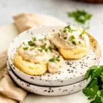 Two biscuits and gravy topped with creamy sauce and garnished with fresh parsley are served on a white speckled plate, with a beige cloth napkin and a sprig of parsley beside them.