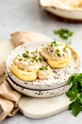 Two biscuits and gravy topped with creamy sauce and garnished with fresh parsley are served on a white speckled plate, with a beige cloth napkin and a sprig of parsley beside them.