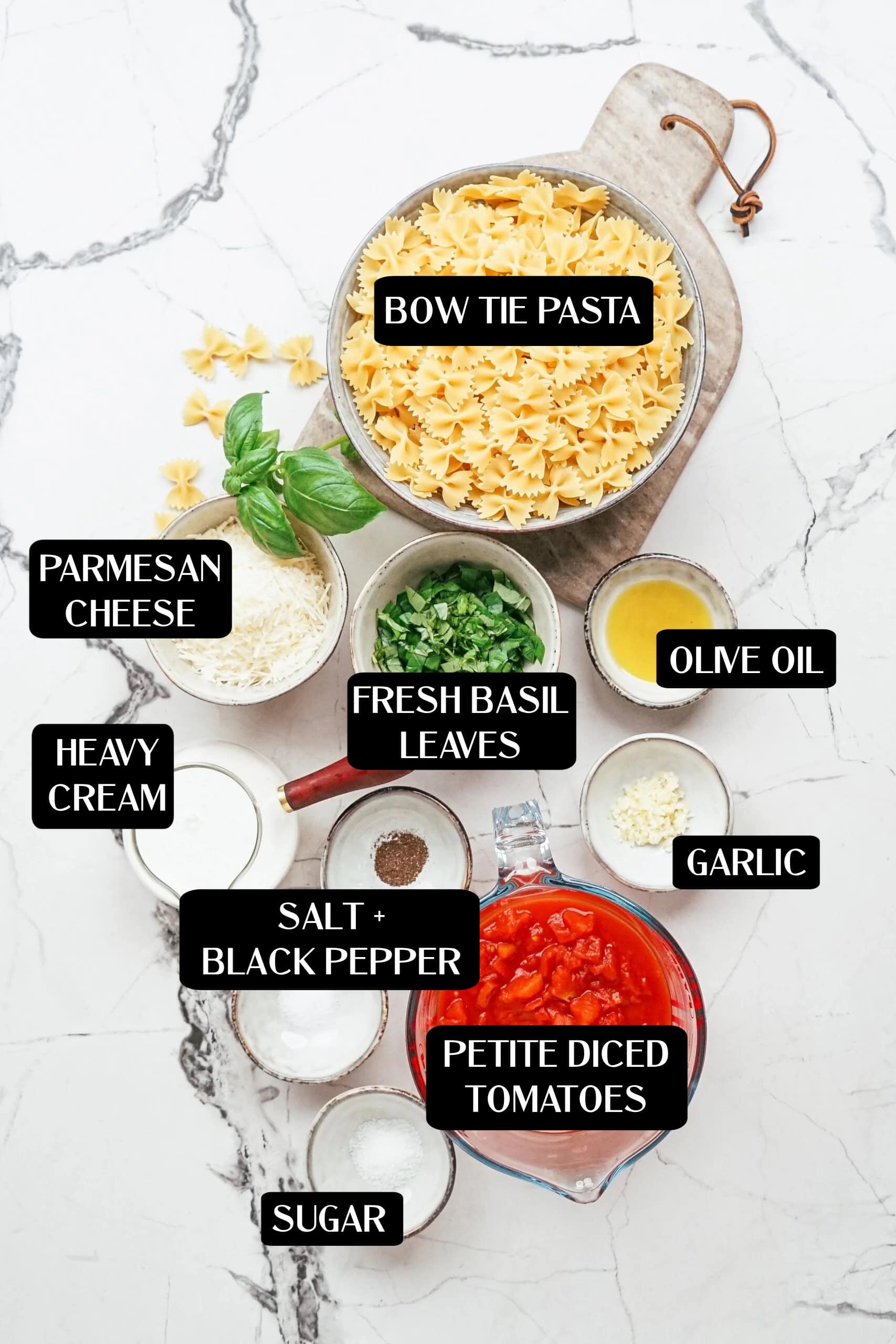 The ingredients for the pasta dish are laid out on a marble surface and feature bow tie pasta, diced tomatoes, parmesan cheese, cream, basil, olive oil, garlic, salt, pepper, and sugar.