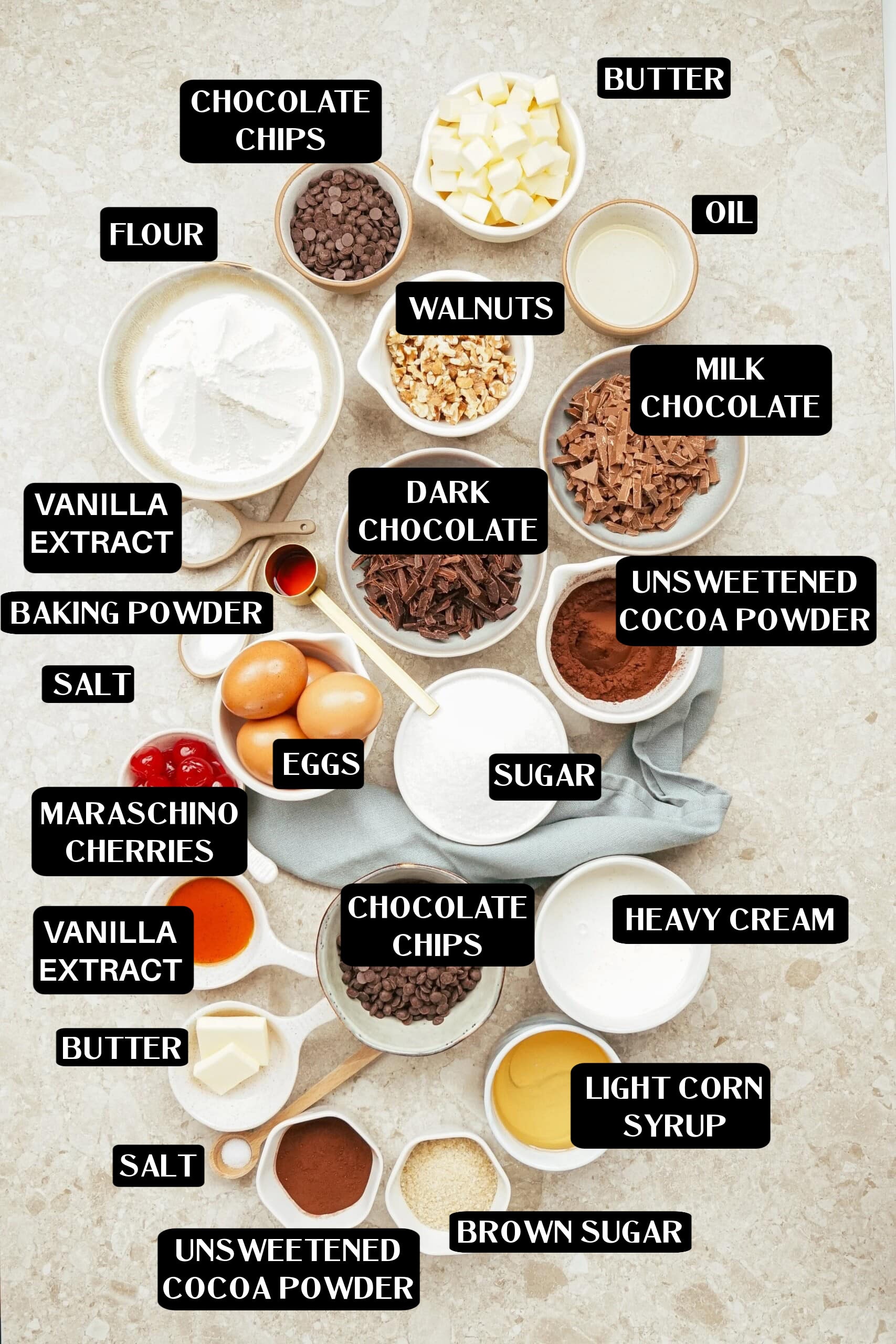 A variety of baking ingredients, each labeled, are arranged on a countertop—perfect for making a delicious Hot Fudge Brownie recipe. Items include flour, cocoa powders, sugars, eggs, butter, oils, nuts, chocolate, cherries, vanilla, and cream.