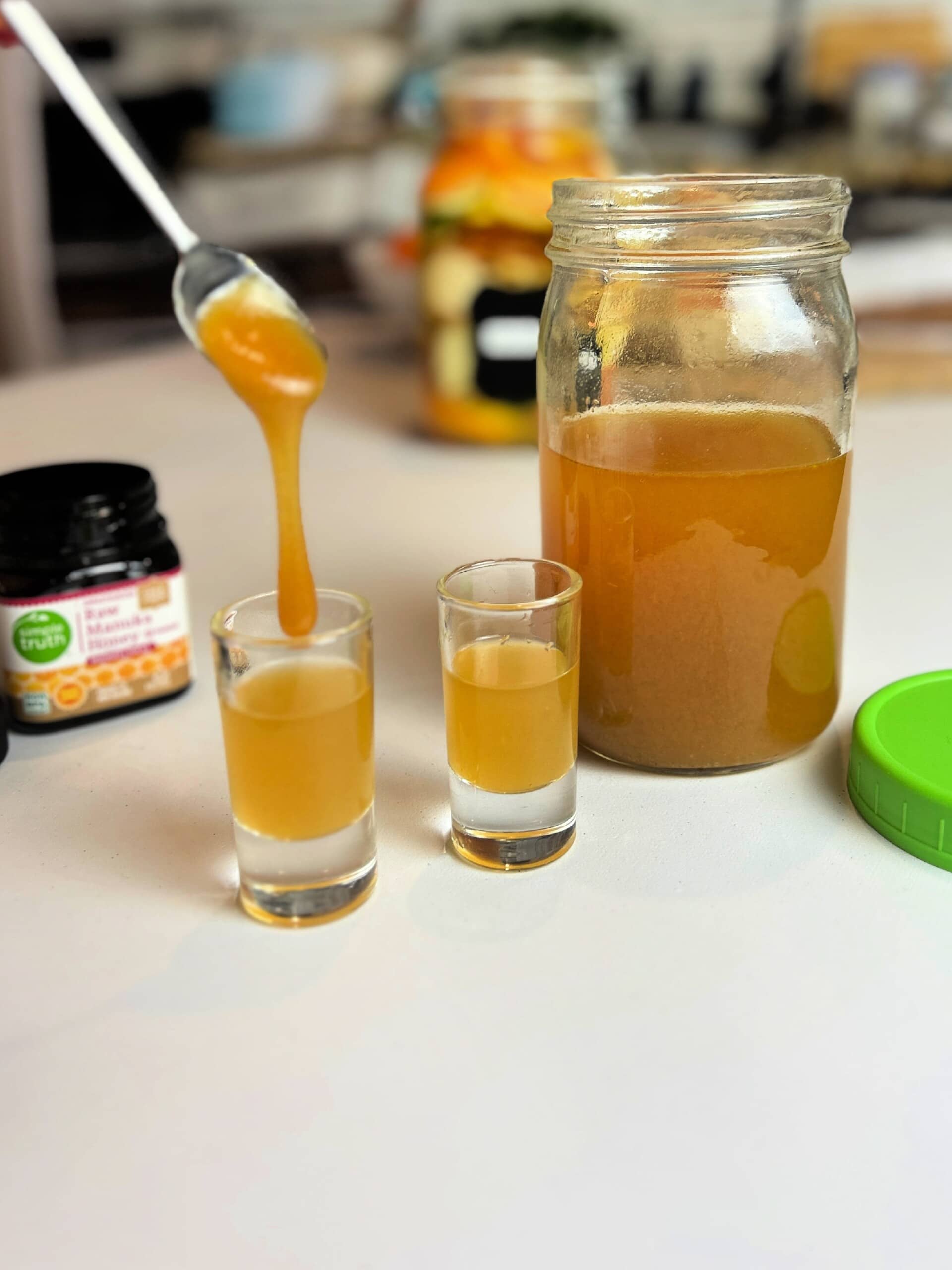 Fire Cider in jar and shot glasses on counter with honey pouring into one glass from spoon