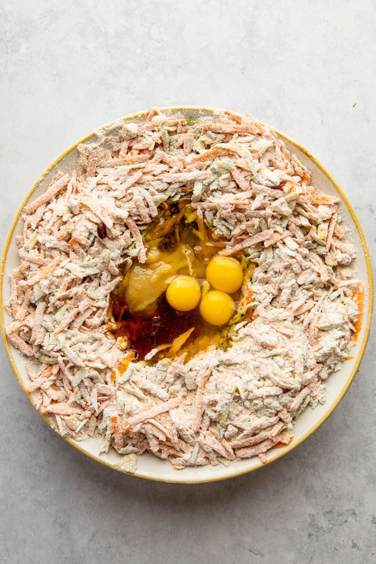 A large bowl containing shredded vegetables and flour, ready to become Morning Glory Muffins, surrounded by three raw eggs and other liquid ingredients in the center.