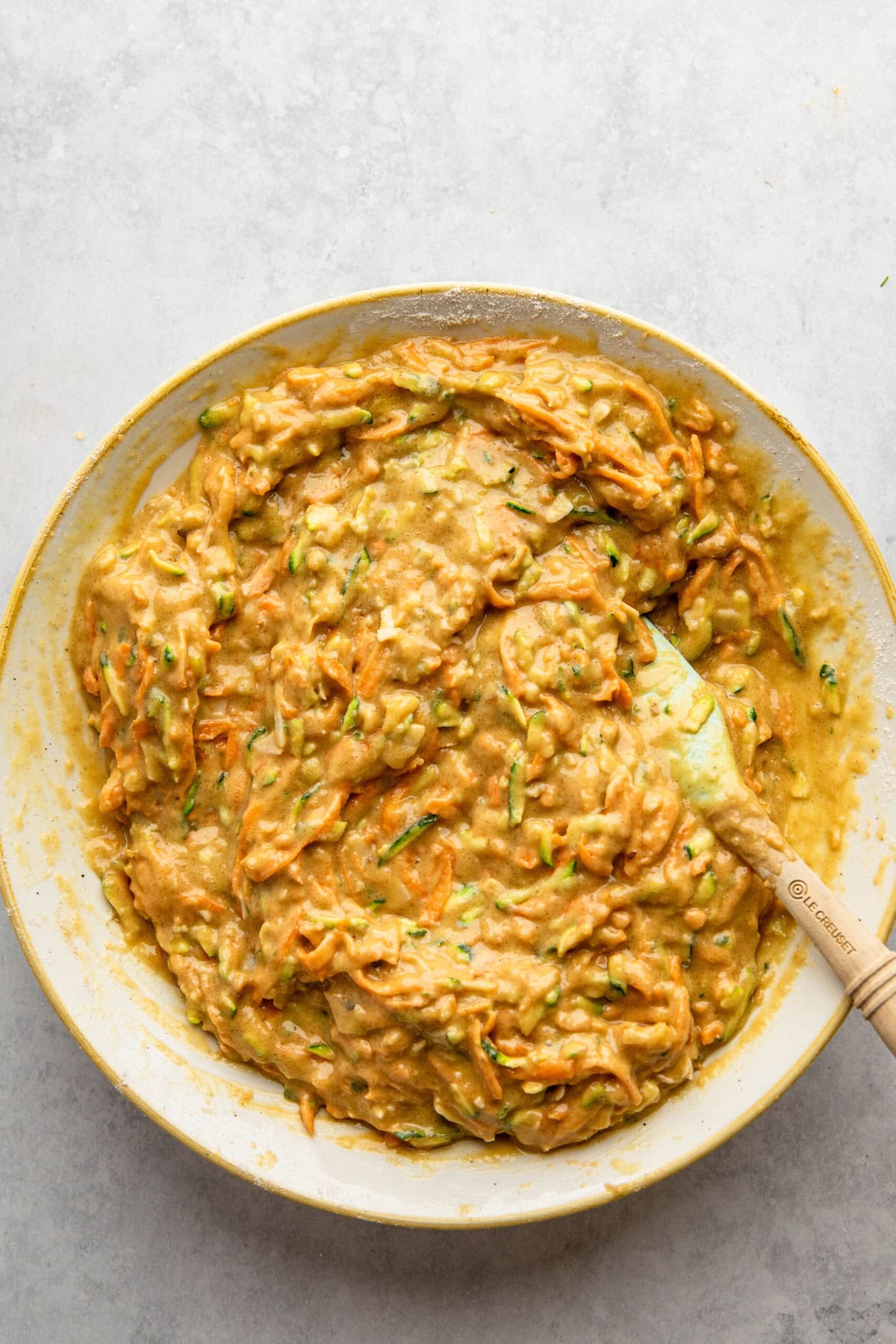 A bowl of zucchini bread batter mixed with a spatula, with visible shredded zucchini and carrots, reminiscent of the wholesome ingredients found in Morning Glory Muffins.