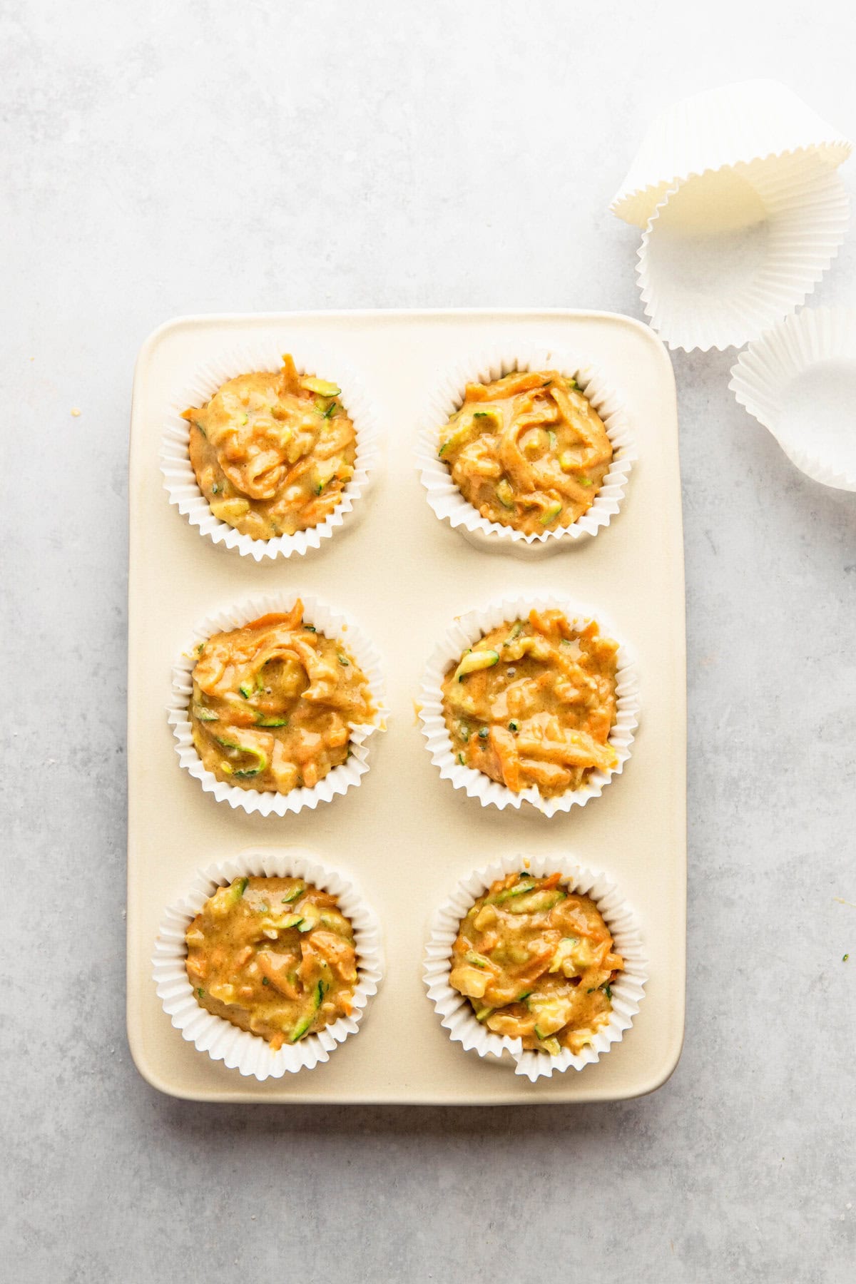 A beige rectangular tray holds six Morning Glory Muffins batter-filled paper liners, ready for baking. Additional empty paper liners are placed to the right on a light gray surface.