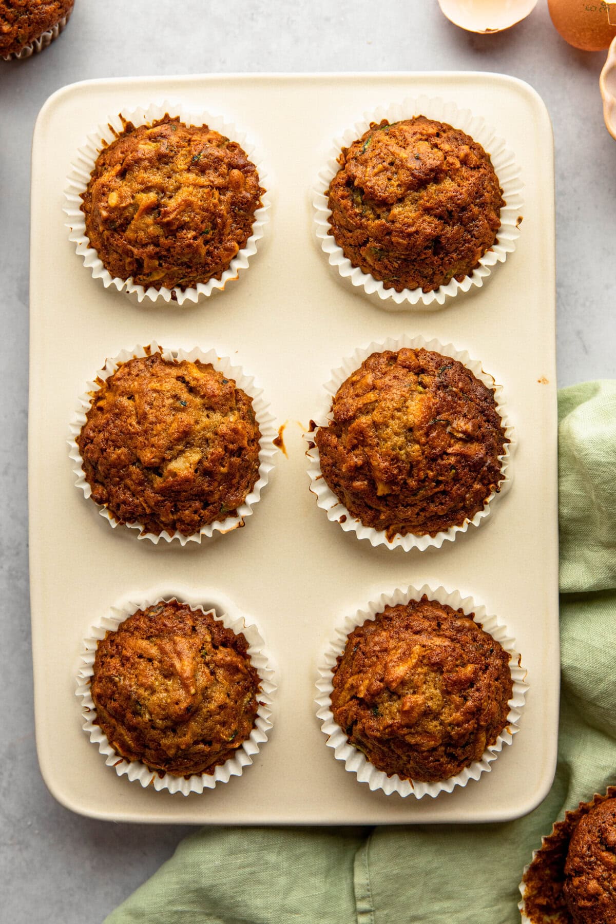 A baking tray with six freshly baked Morning Glory Muffins in white paper liners. A green cloth and broken eggshells are visible around the tray.