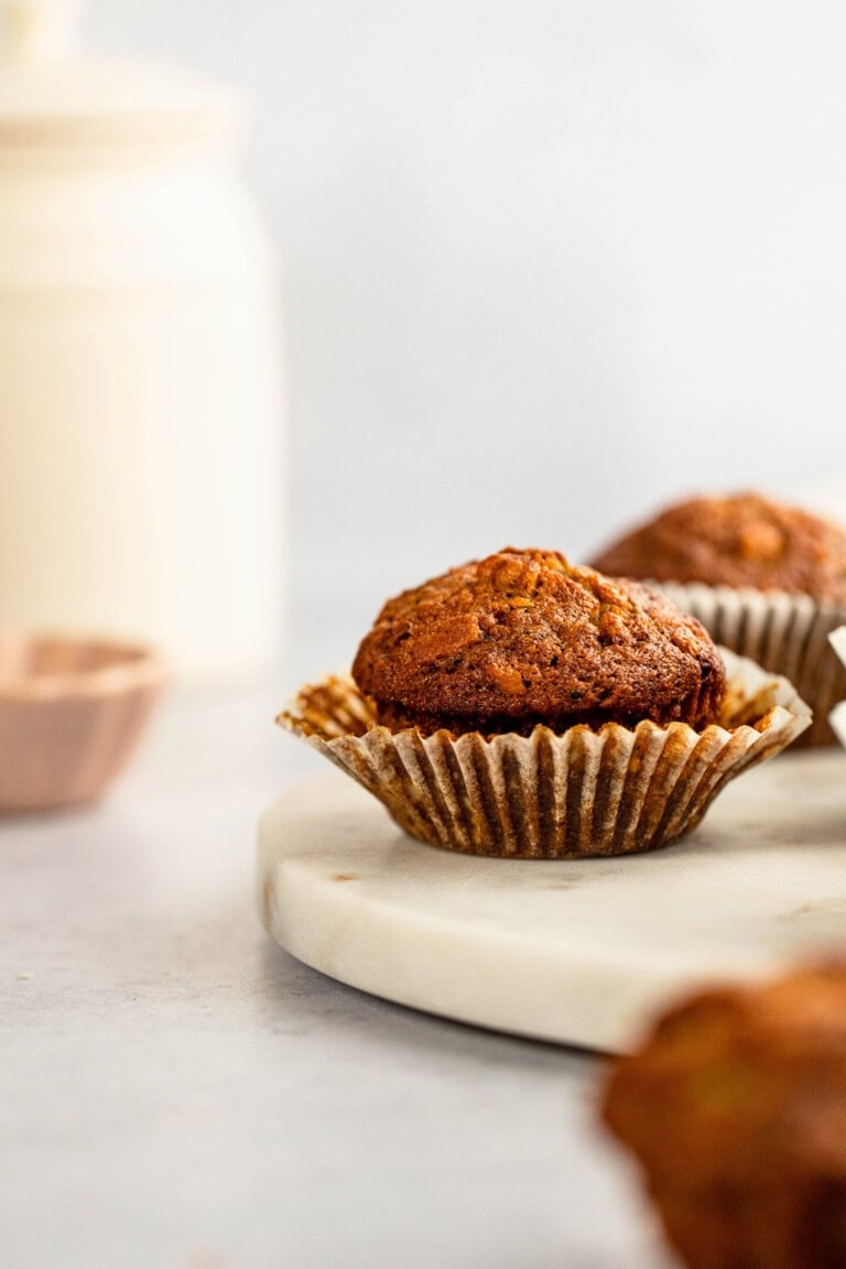 Close-up of two Morning Glory Muffins in paper liners on a white marble surface, with a blurry background showing a white container and a measuring cup.