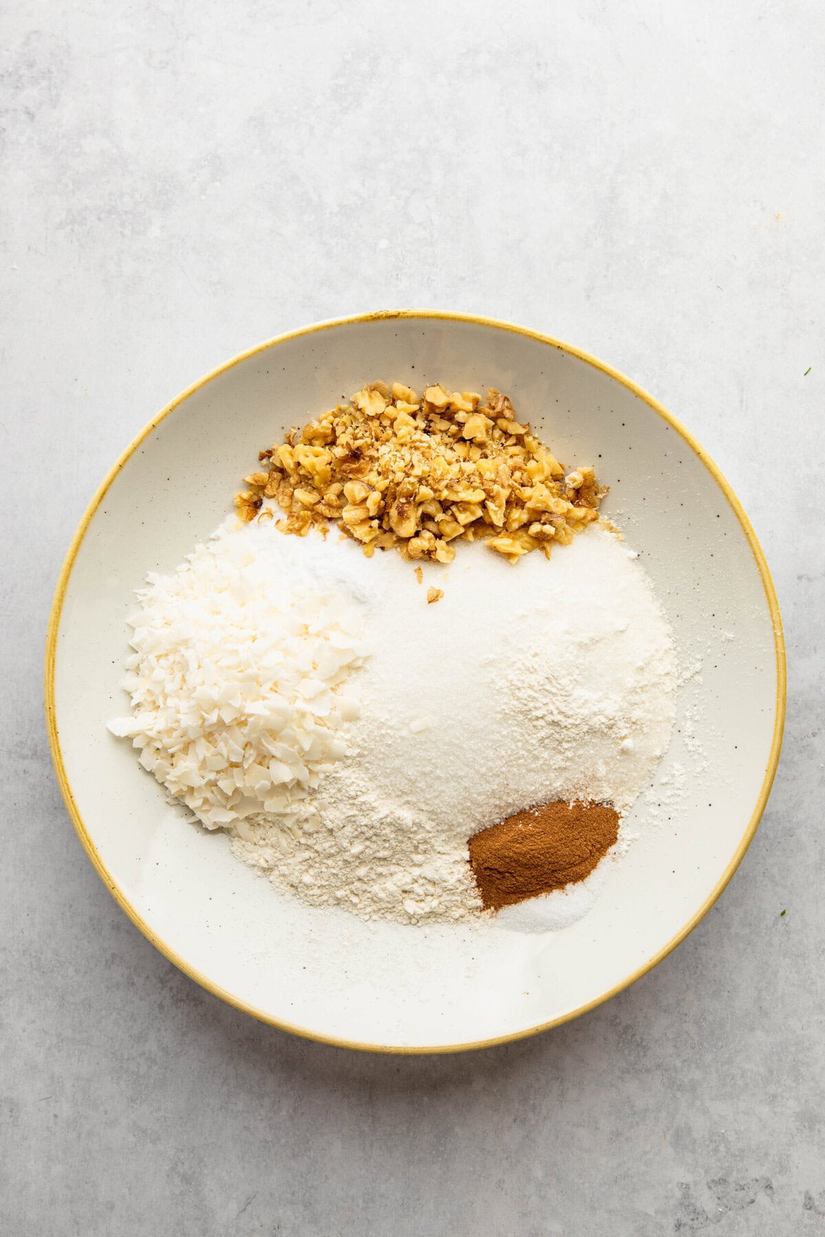 A plate with four piles of ingredients: chopped nuts, shredded coconut, flour, and ground cinnamon on a light gray background—perfect for baking delicious Morning Glory Muffins.