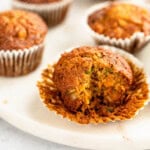 A close-up of several Morning Glory muffins, with one muffin partially unwrapped and bitten, resting on a marble surface. Crumbs are scattered nearby.