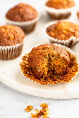 A close-up of several Morning Glory muffins, with one muffin partially unwrapped and bitten, resting on a marble surface. Crumbs are scattered nearby.