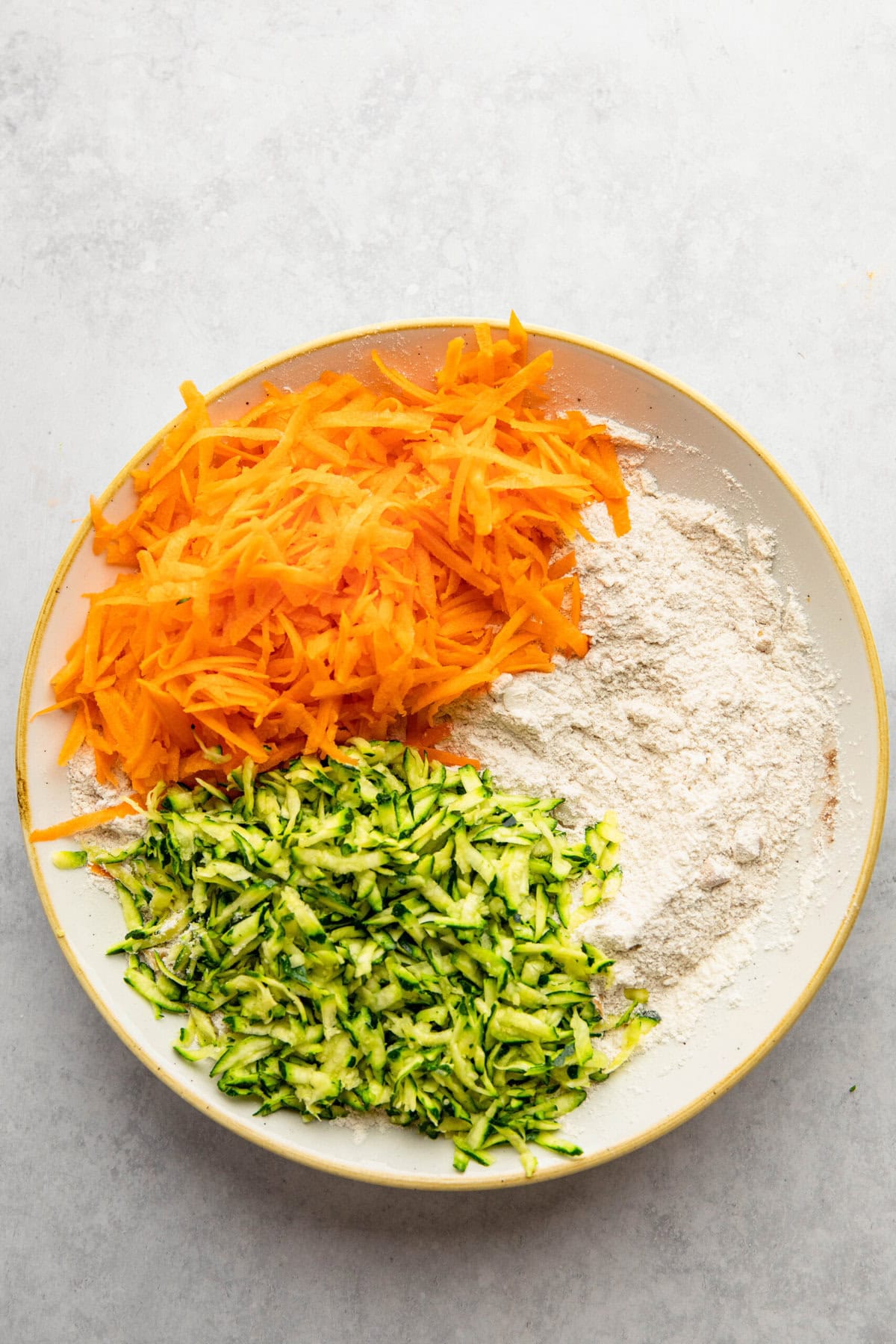 A plate with shredded carrots, shredded zucchini, and a pile of flour on a light gray surface—essentials for fresh Morning Glory Muffins.