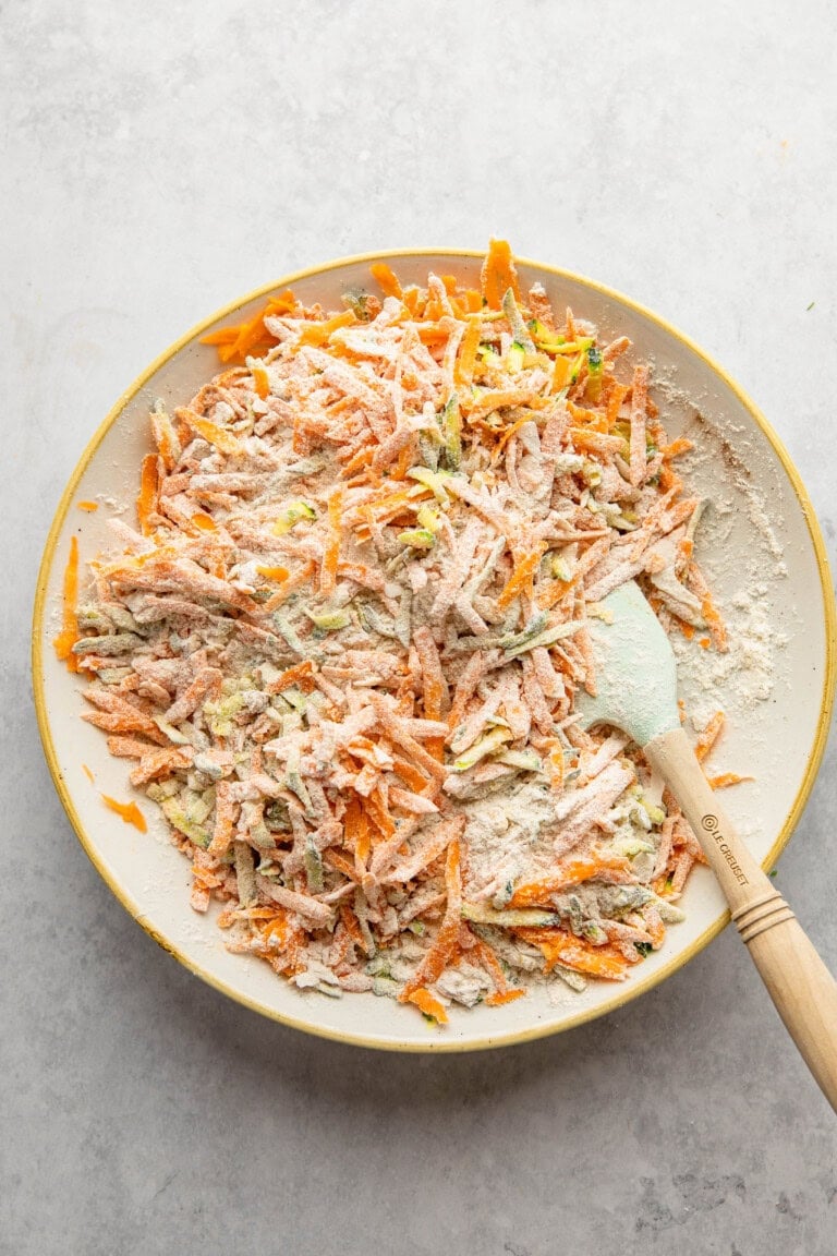 A plate contains shredded vegetables, primarily carrots, mixed with flour, reminiscent of the ingredients for Morning Glory Muffins. A mixing spatula rests on the right side of the plate.