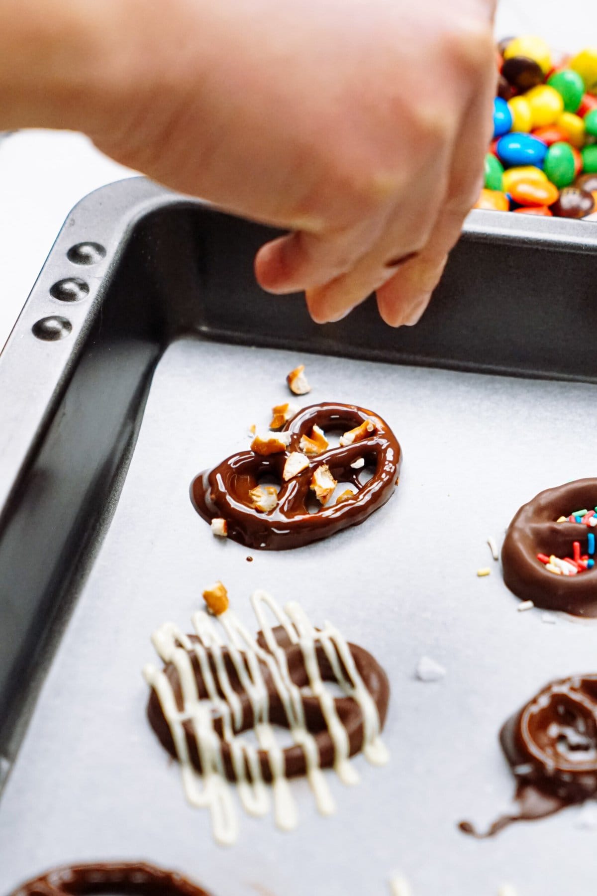 A person is putting crushed pretzel pieces on a chocolate covrered pretzel on a baking sheet.
