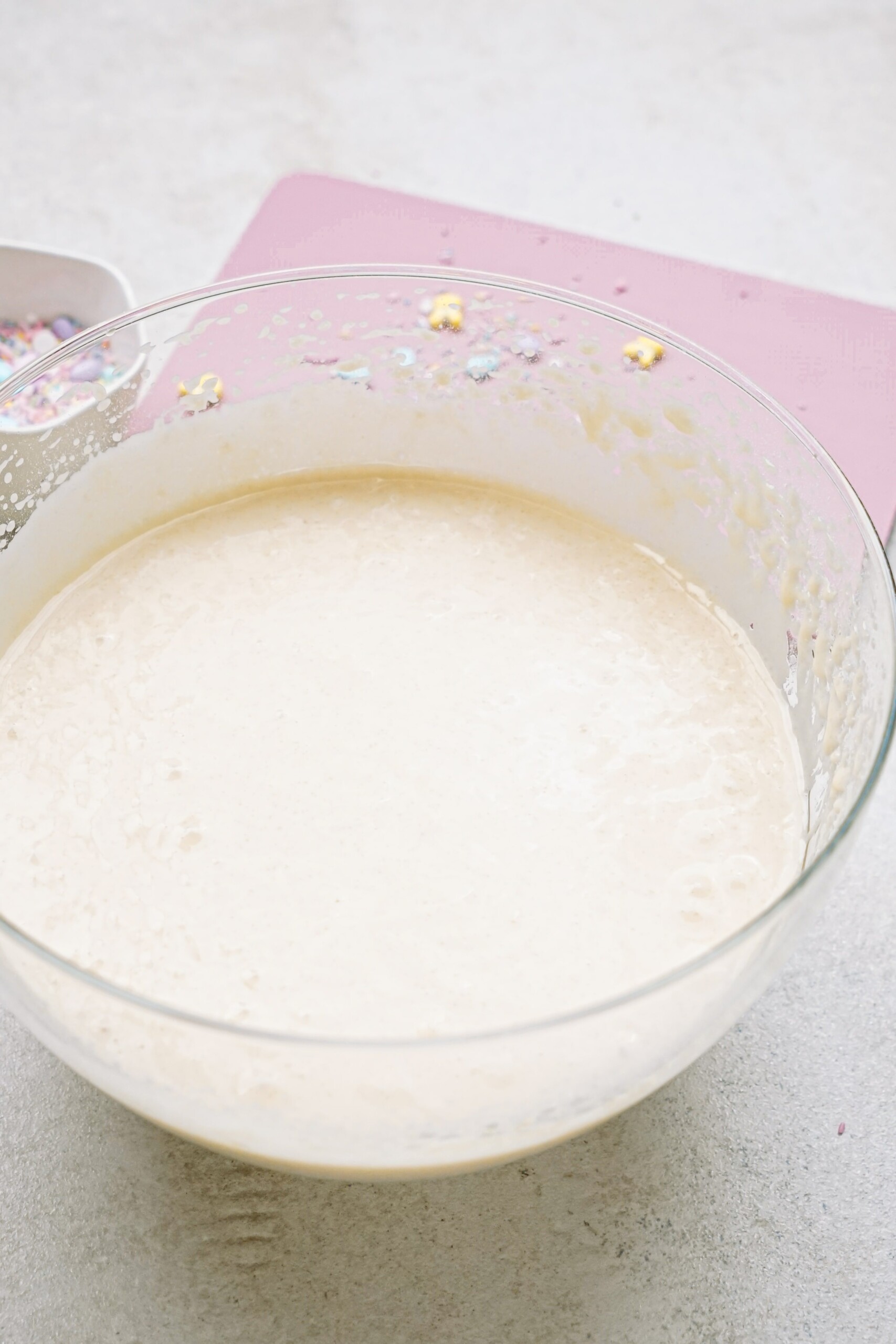 A bowl of cake batter on a pink mat, with visible air bubbles and a whisk partially in view.