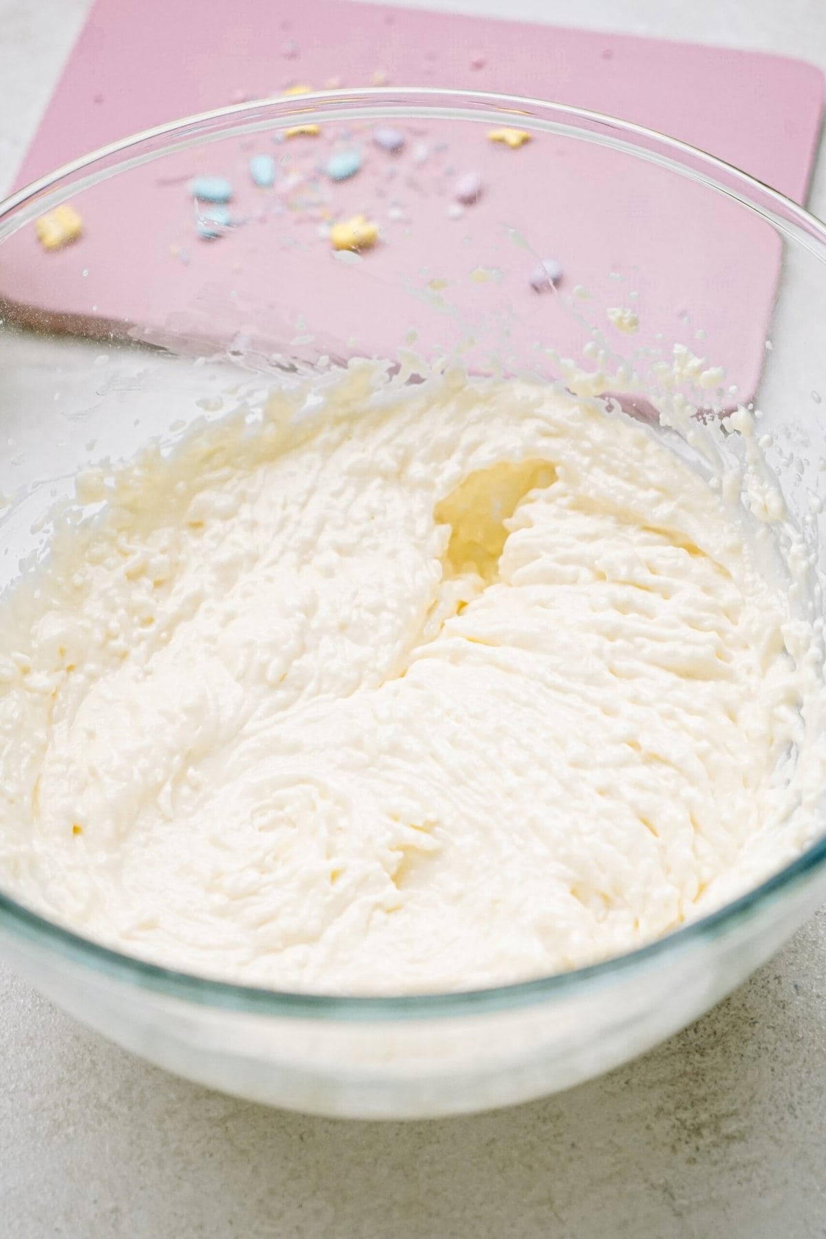 Bowl of frosting on a kitchen counter with a pink spatula in the background.
