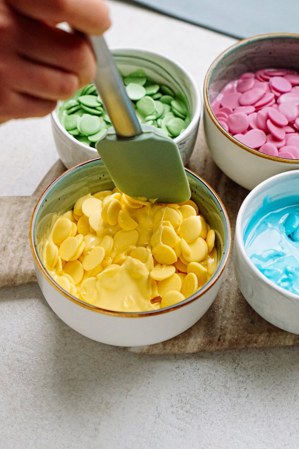 Melting yellow candy melts in a bowl with a spatula, surrounded by other bowls of various colored candy melts.