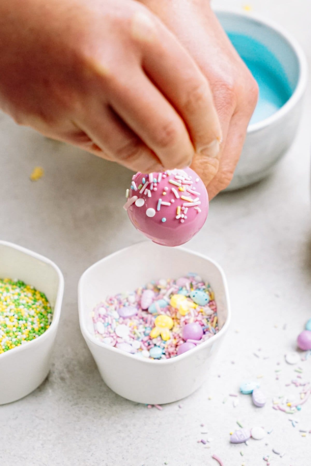 Decorating a pink cake pop with sprinkles over a bowl.