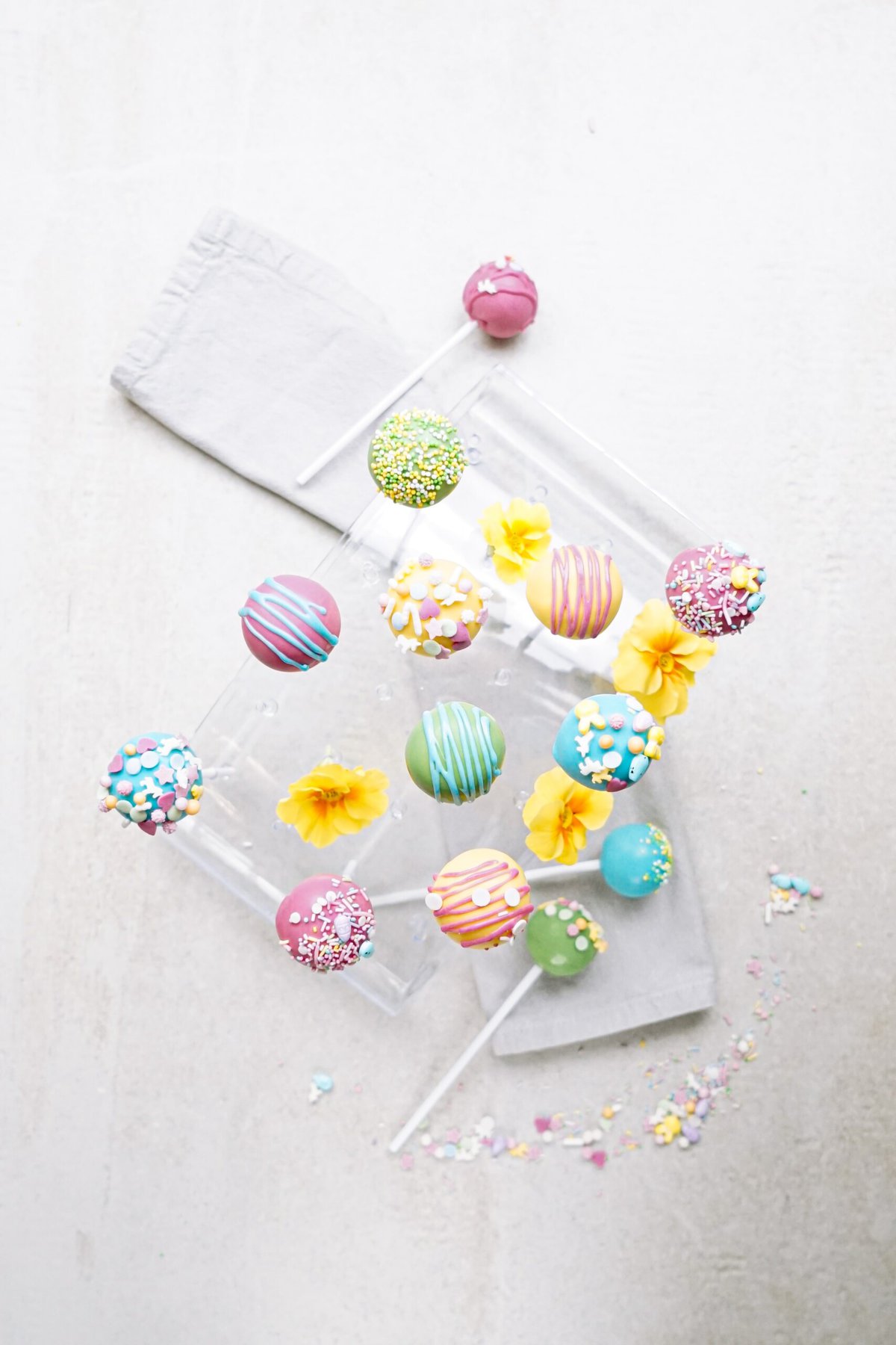 Assorted colorful cake pops with flower decorations and sprinkles on a light background.
