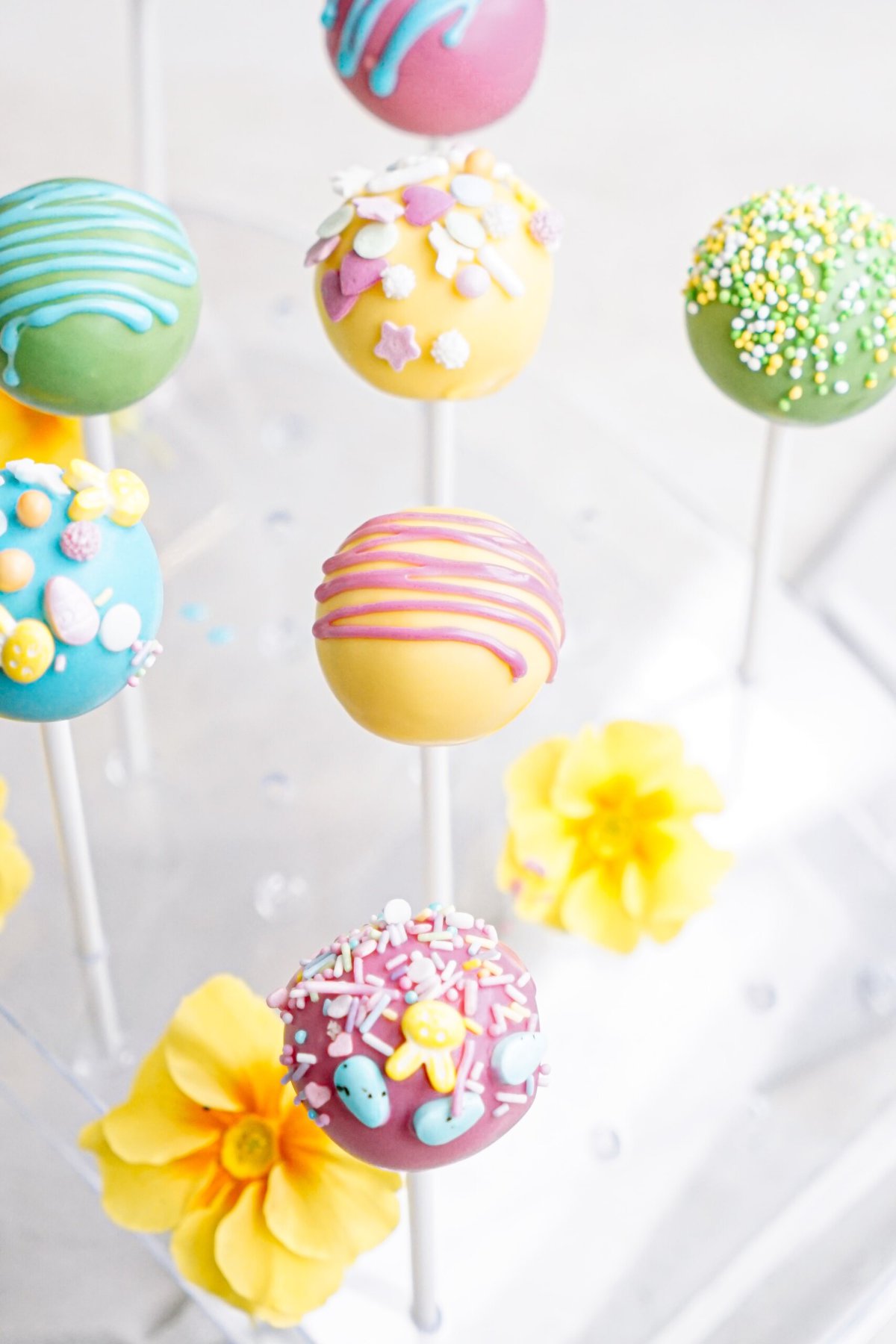 Assorted cake pops adorned with colorful icing and sprinkles displayed on a stand with yellow flowers.