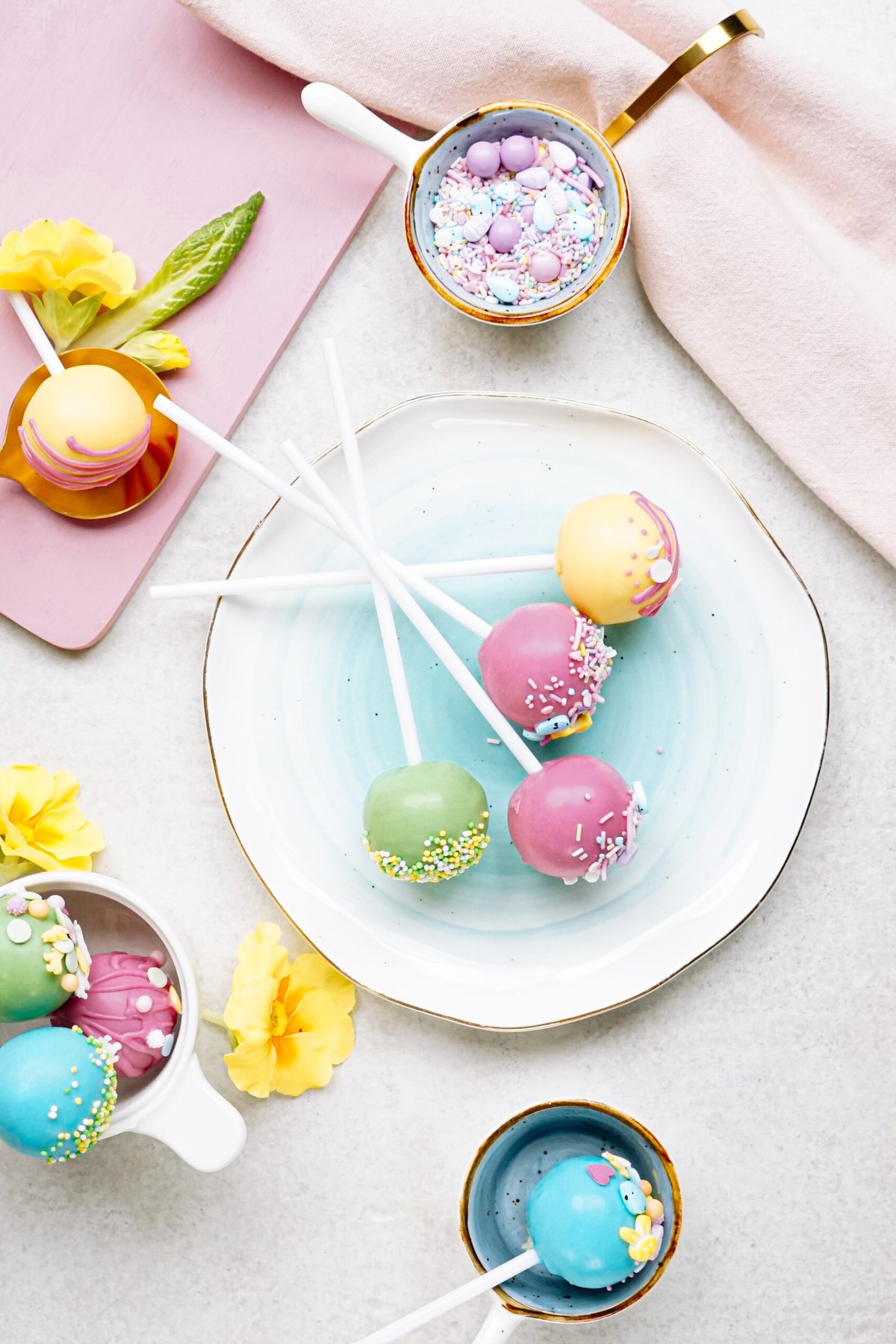 Colorful cake pops decorated with sprinkles, presented on a pastel dish alongside spring flowers and decorative eggs.