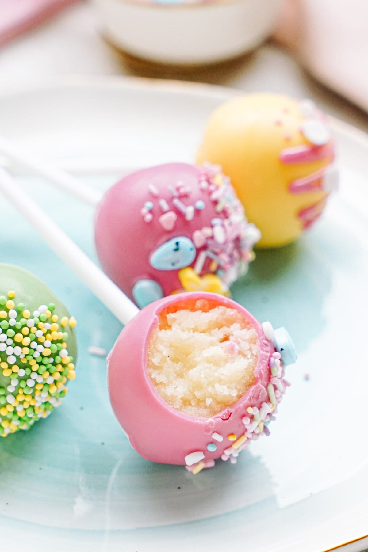 Colorful cake pops with bite taken out of one, decorated with sprinkles, displayed on a plate.