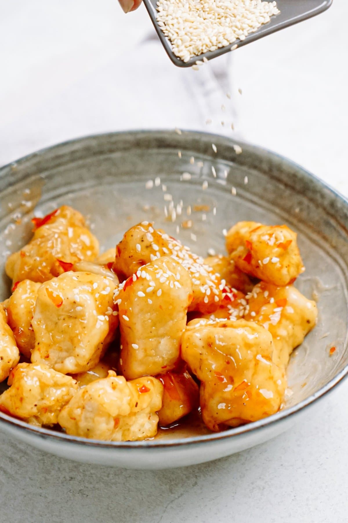 A person is sprinkling sesame seeds on chicken in a bowl.