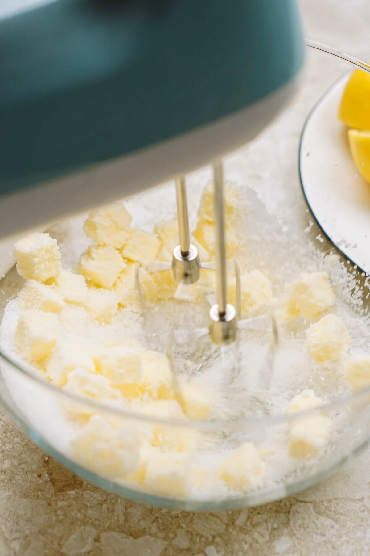 An electric mixer beating butter and sugar in a glass bowl, with lemon wedges in the background.