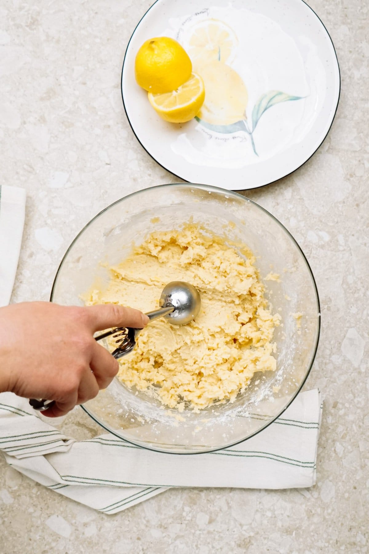 A person using an ice cream scoop to portion cookie dough into a bowl, with sliced lemons on a plate nearby.