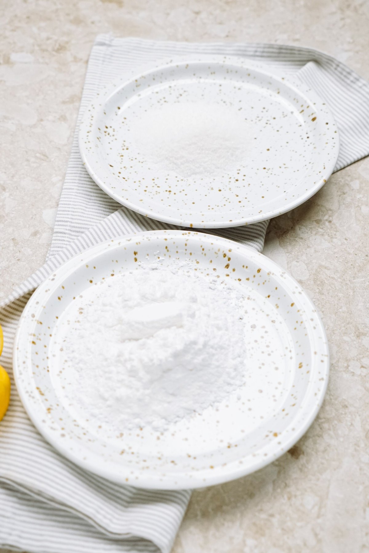 Two white, speckled plates with powdered sugar on a marble surface, one plate partially covering a striped cloth napkin.