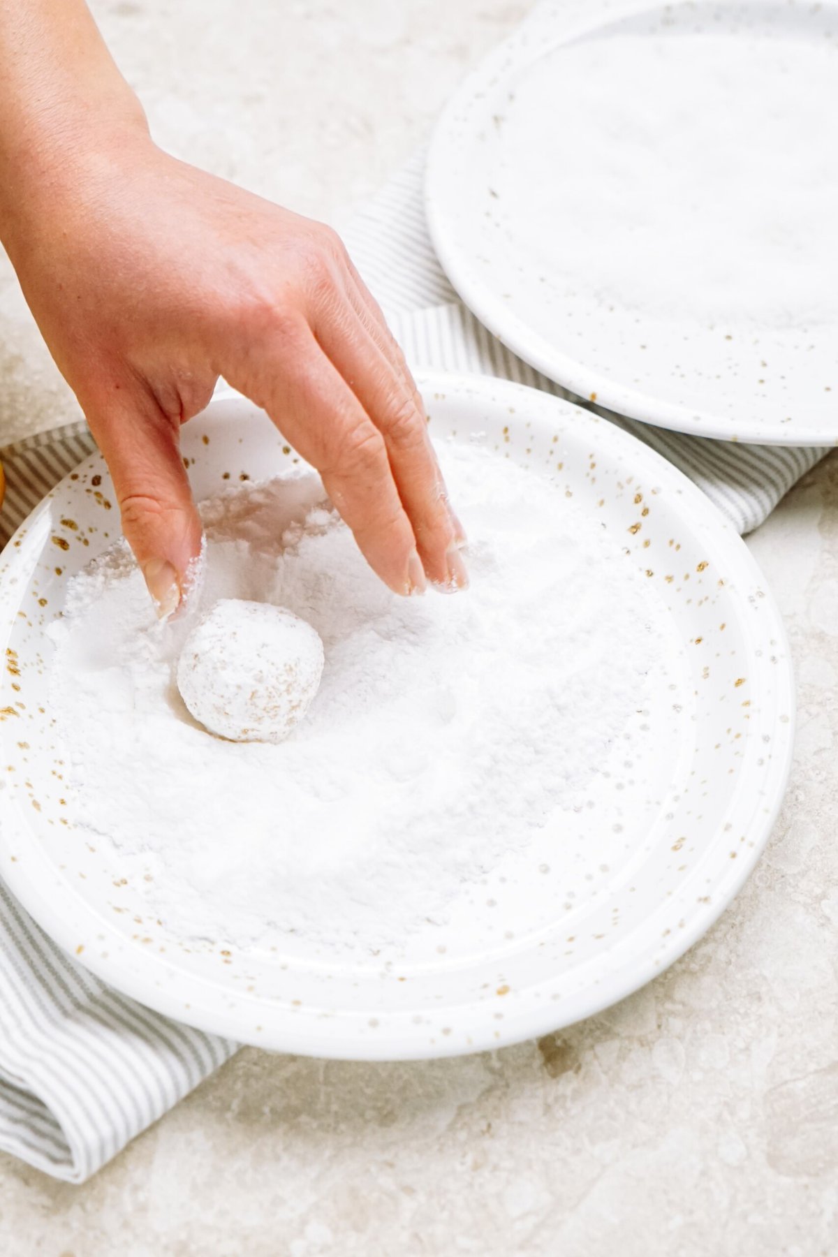 A person's hand coating a small round dough ball in powdered sugar on a white plate.