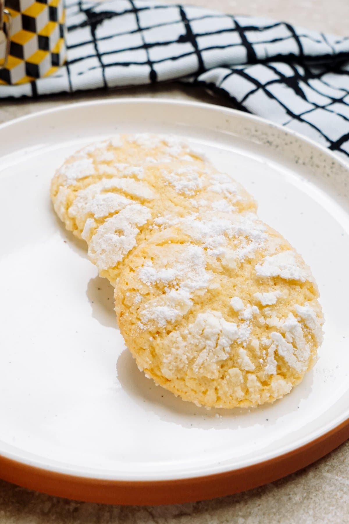 Three crinkle lemon cookies with powdered sugar on a white plate with a terracotta rim, accompanied by a black and white checked cloth.